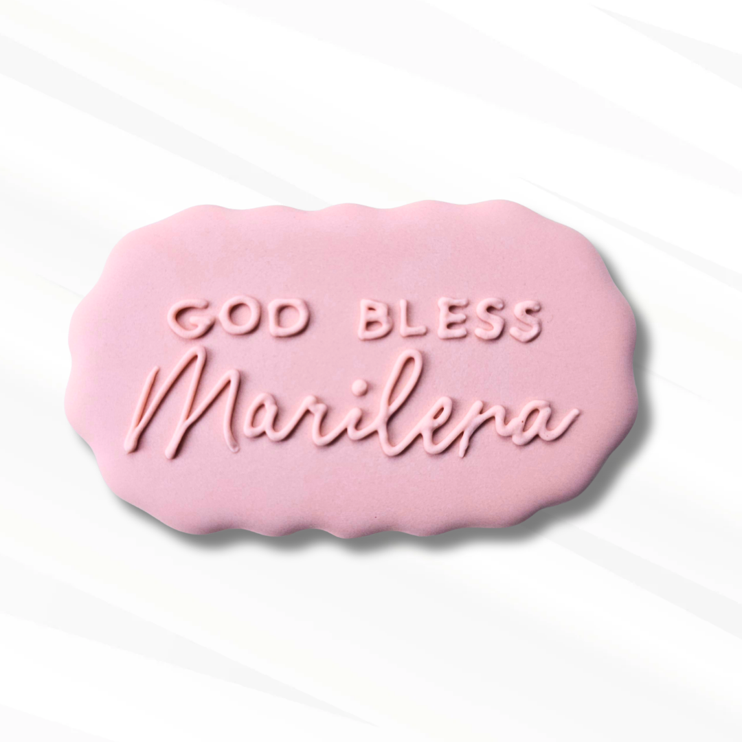 A pink scalloped, oval-shaped sugar cookie that has God bless Marilena piped onto it in pink icing.