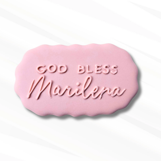 A pink scalloped, oval-shaped sugar cookie that has God bless Marilena piped onto it in pink icing.