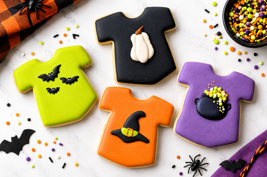 Four Halloween-themed t-shirt-shaped sugar cookies decorated with colorful Halloween royal icing transfers, arranged on a white marble surface with sprinkles, plastic spiders, and a black bowl of sprinkles